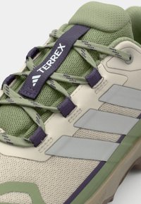 Beige sportsko med nettstoff, grønne detaljer og lilla snøring. Har tre hvite striper og polstret tunge for komfort.