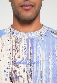 Monivärinen collegepaita sinisenä, kermanvärisenä ja ruskeana. Koristeena abstrakteja kuvioita, resorikaulus ja kirjailtu logo rinnassa. Pehmeä pinta.