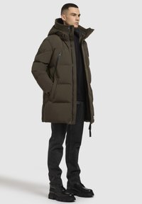 Puffervest i mørk olivengrøn med hætte, lynlåslommer og kviltet design. Båret over en sort top og parret med mørke jeans og kraftige støvler.