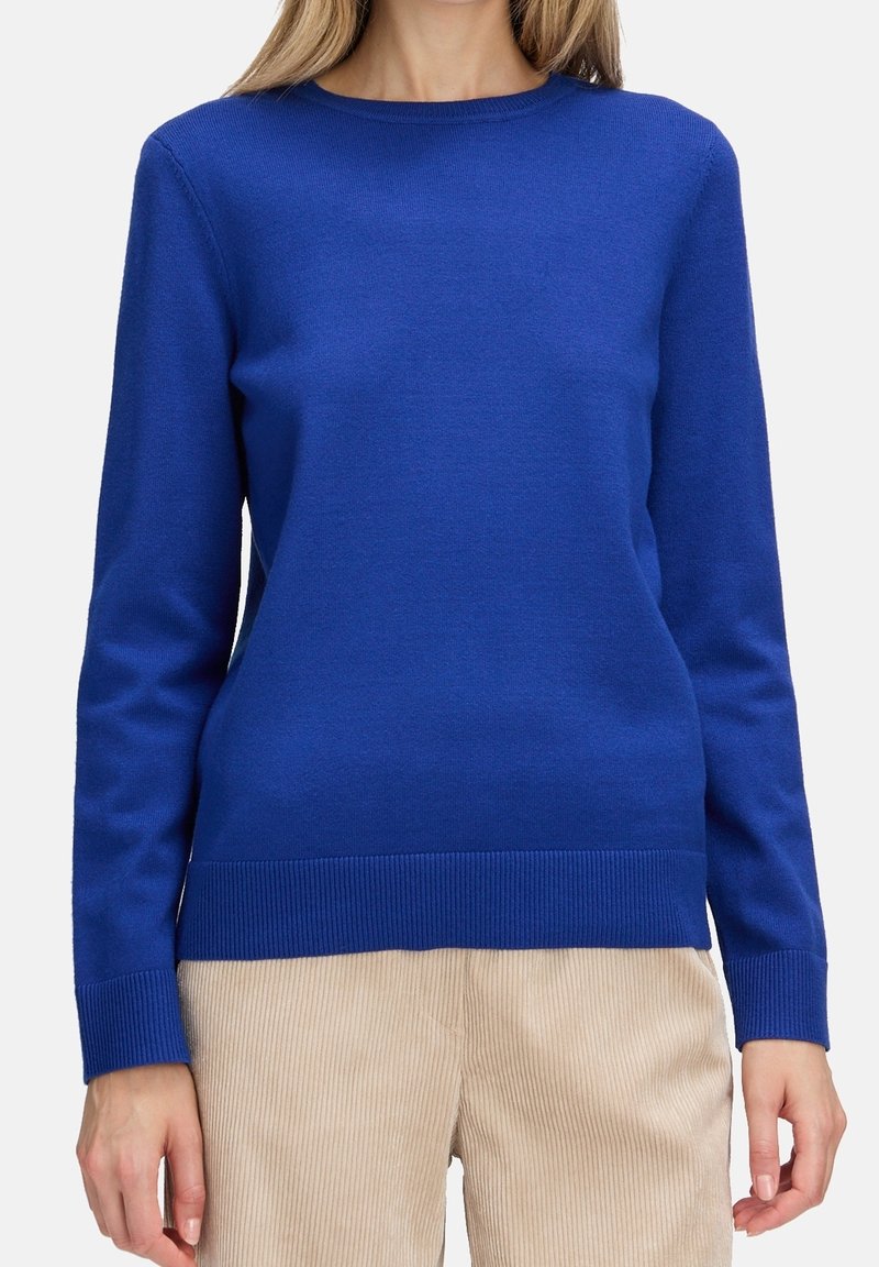 Femme portant un pull bleu roi uni à manches longues associé à un pantalon beige en velours côtelé, debout sur un fond blanc.