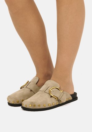 Zoccoli beige in camoscio senza lacci con borchie dorate e grande fibbia dorata, indossati a piedi nudi.