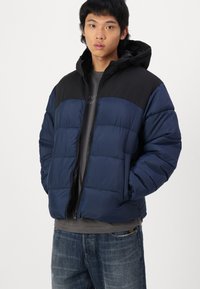 Navy blue padded jacket with black upper, zip closure, and attached hood. Features a quilted texture and elastic cuffs. Worn over a grey shirt.