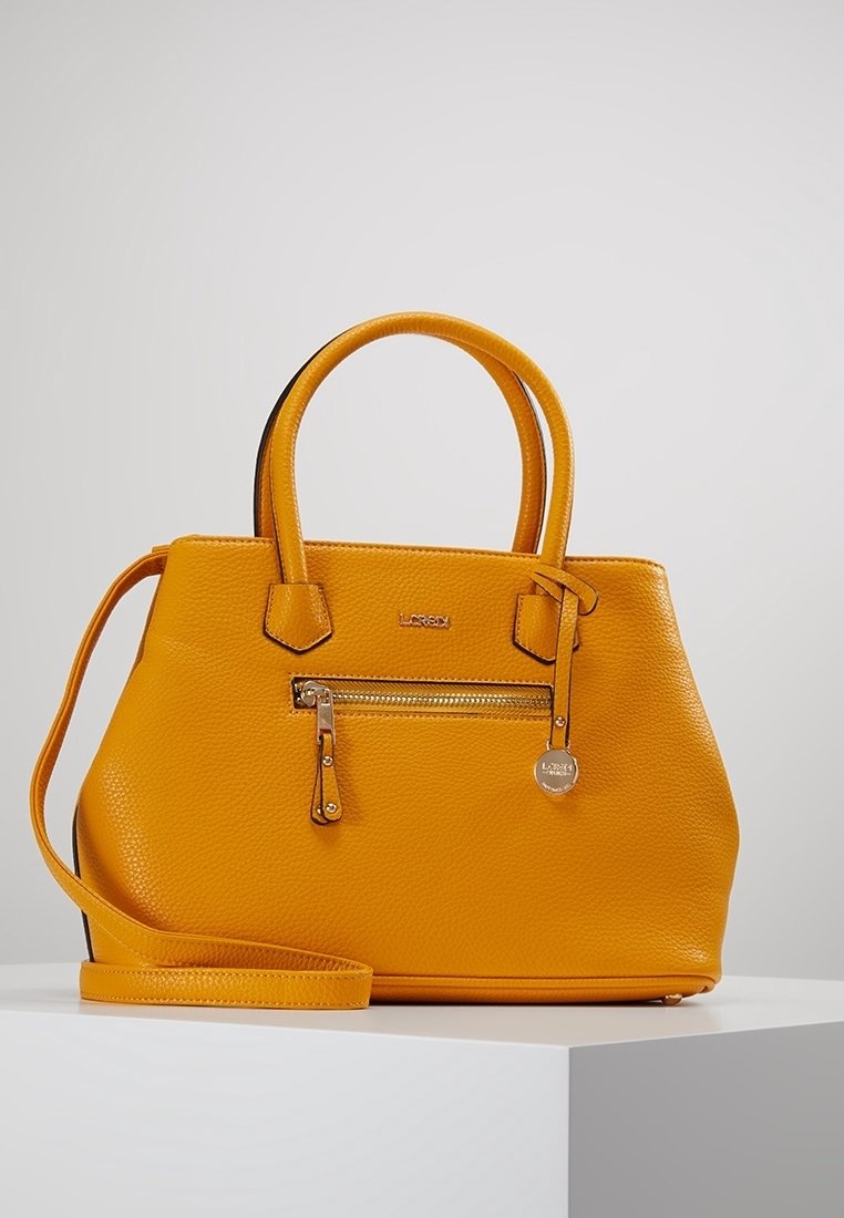 Sac à main en cuir jaune texturé avec deux poignées, poche avant à fermeture éclair, pendentif métallique de marque, et bandoulière amovible sur un piédestal blanc.