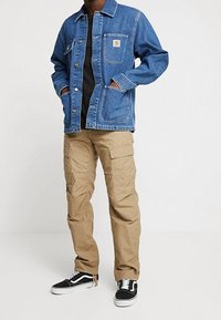 Farkkutakki napilliseen suljettuun, rinnantaskulla ja sivutaskuilla, yhdistettynä khakinvärisiin cargo-housuihin, joissa on sivulla kuormitustaskut, sekä mustiin lenkkareihin.