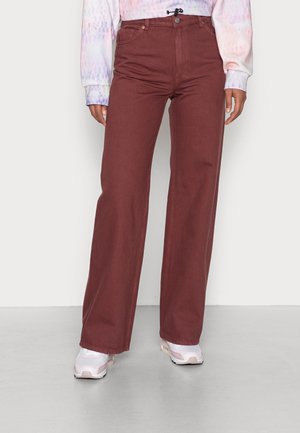 Personne portant un pantalon large bordeaux et des baskets blanches, debout devant un fond clair uni.