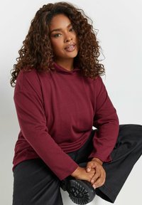 Felpa bordò con maniche lunghe, vestibilità rilassata e colletto regolabile con lacci. Indossata con pantaloni neri; tessuto morbido e design casual.