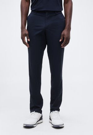 Moški, oblečen v oprijete temno modre hlače in bele golf čevlje, stoji ob enostavni svetli podlagi, z rokami sproščeno ob straneh.