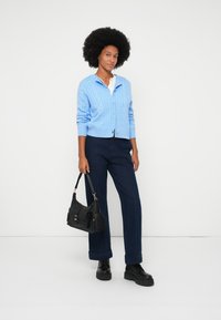 Gai zila adīta jakas, balta apkakles krekla, tumši bikses, melna rokassoma un melni masīvi zābaki, fona laikā uz baltas virsmas.