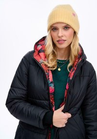 Čierna pufová bunda s kvetinovou podšívkou, žltá pletená čiapka, zelený sveter pod ňou, so zipsom vpredu a rebrovanými manžetami.