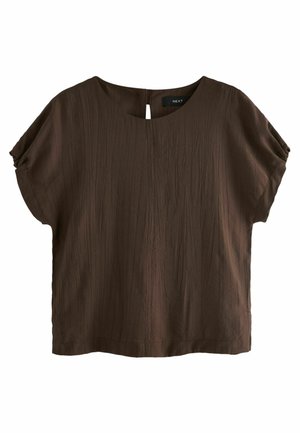 Vrouw die een bruin gestructureerd kortemouwshirt draagt, gecombineerd met een luipaardprint broek, staand tegen een beige achtergrond.