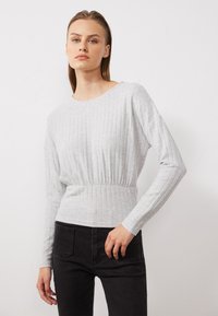 Lichtgrijze longsleeve top met geribbelde textuur en een geplooide taille, gecombineerd met zwarte jeans. Neutrale kleur en eenvoudig ontwerp.