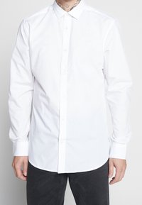 Camicia bianca con bottoni realizzata in tessuto misto cotone, con colletto classico, maniche lunghe e orlo curvo. Senza motivi né dettagli.