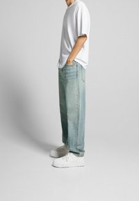 Persona vistiendo una camiseta blanca holgada, jeans claros y anchos, y zapatillas blancas, de pie con las manos en los bolsillos, contra un fondo claro y liso.