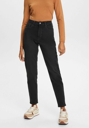 Person trägt schwarze High-Waist Straight-Leg Jeans, beigen Langarmpullover und weiße-braune Sneakers, steht vor einem schlichten Hintergrund.