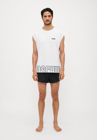 White sleeveless t-shirt with black "MOSCHINO" logo and text at the hem, paired with black shorts. Model stands barefoot against a white background.
