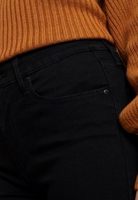 Pantalones de mezclilla negros con un bolsillo frontal visible, combinados con un suéter de punto acanalado color ámbar. Textura suave, diseño ajustado.