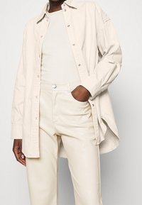 Camicia over-size beige chiaro in cotone, con chiusura a bottoni, abbinata a pantaloni in ecopelle crema a vita alta.