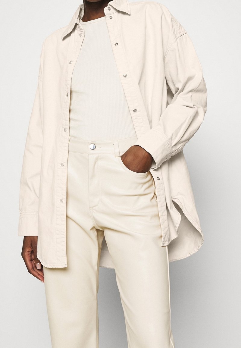 Camicia over-size beige chiaro in cotone, con chiusura a bottoni, abbinata a pantaloni in ecopelle crema a vita alta.