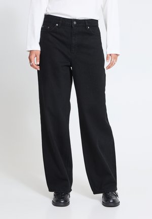 Personne portant un jean noir à jambes larges, un haut blanc à manches longues, des chaussures noires et des bagues argentées aux deux mains visibles de la taille vers le bas.