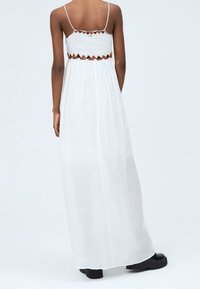 Weißes Maxikleid mit dünnen Trägern, das mit einem bunten bestickten Saum in der Taille versehen ist. Leichter, fließender Stoff mit einer sanften Textur.