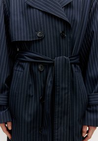 Mörkblå randig trenchcoat med dubbelknäppt design, stora knappar och knytbart midjeband för justerbar passform.