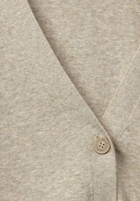 Beige neulecardigan, jossa on pehmeä tekstuuri ja V-kaula-aukko. Siinä on yksi pyöreä nappidetaili. Kangas on kevyt ja sileä.