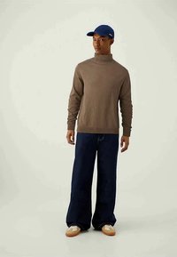 Suéter de gola alta castanho em malha, boné azul-marinho, jeans escuros largos e ténis bege com detalhes brancos. Roupa casual com um ajuste descontraído.