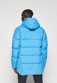Parka albastră pufoasă cu glugă, având detalii cu cusături orizontale și un logo negru pe spate, fabricată dintr-un material neted, rezistent la intemperii.