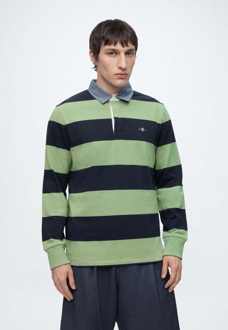 Homme debout devant un fond uni, portant un polo de rugby à rayures vertes et bleu marine avec un col bleu et un pantalon foncé ample.