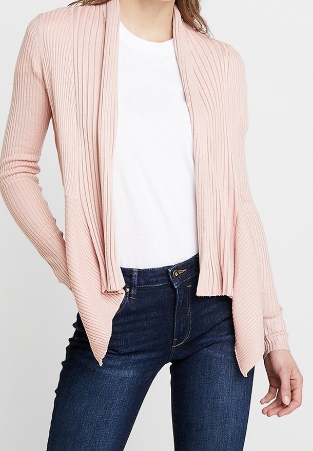 Frau, die eine hellrosa gerippte offene Strickjacke über einem weißen T-Shirt und dunklen, hoch taillierten Jeans trägt, steht mit einer Hand in der Tasche.