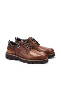 Zapatos de cuero marrón con cordones, puntera redonda, suelas de goma marrón texturizadas y cordones oscuros que contrastan, con detalles decorativos de costura.