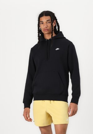 Mladi moški s spetimi lasmi, oblečen v črno Nike kapucarico in rumene Nike kratke hlače, stoji pred enobarvnim belim ozadjem.