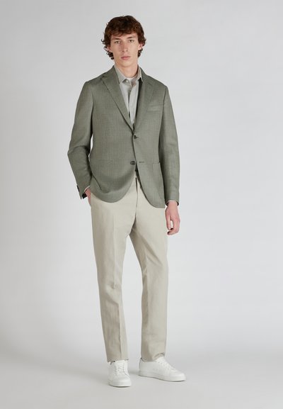 Blazer cintré vert clair en tissu texturé, fermeture à un bouton et poches plaquées, associé à un pantalon beige clair et des baskets blanches.