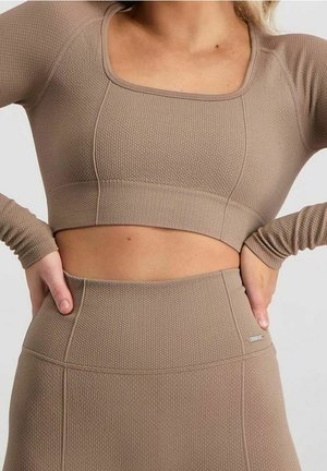 Woman wearing a textured beige long-sleeve crop top and high-waisted leggings, hands resting on hips against a plain background.