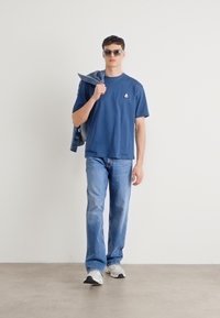 Blå t-shirt med kortærmer og logo, lyseblå denim jeans og hvide sneakers. Model holder en denimjakke over skulderen.