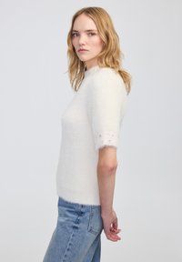 Witte, fluffy trui met korte mouwen en een hoge kraag. Beschikt over getextureerde mouwen versierd met kleine, sprankelende details.
