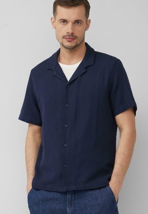 Mann mit heller Haut und kurzen braunen Haaren, der ein dunkelblaues kurzärmeliges Hemd mit Knöpfen über einem weißen T-Shirt trägt, Hände in den Taschen einer blauen Jeans.