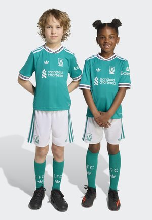 Uniformes de fútbol para niños en color teal con acentos blancos, que incluyen el logo de "Standard Chartered", pantalones cortos y calcetas hasta la rodilla. Botines negros.