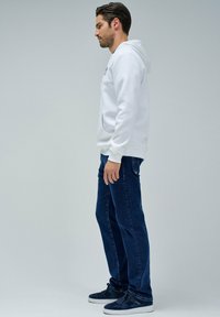 Sudadera blanca con capucha y bolsillo frontal, combinada con unos vaqueros rectos de color azul oscuro y zapatillas de deporte navy. El conjunto es casual y cómodo.