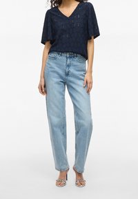 Woman wearing a navy patterned short-sleeve top, light blue high-waisted straight jeans, and white high-heeled sandals with metallic details.