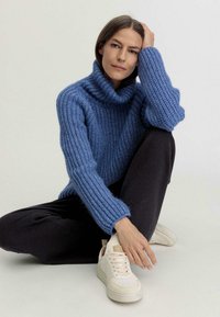 Chunky blauer Strickpullover mit hohem, gerolltem Kragen und gerippter Textur, kombiniert mit schwarzen Hosen und cremefarbenen Sneakers.