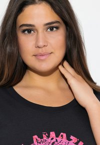 Černé tričko s kulatým výstřihem s živým růžovým grafickým designem. Hladká textura se krátkými rukávy, zdůrazňující ležérní styl.