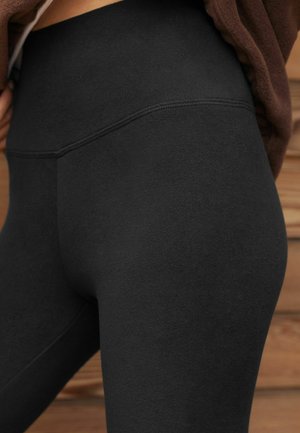 Detailní záběr osoby v černých legínách s vysokým pasem a viditelnými švy, s hnědým topem, stojící na pozadí dřeva.