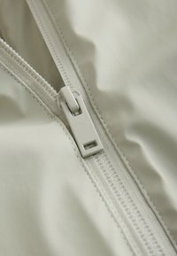 Prim-plan al unui cursor de fermoar alb parțial închis pe o țesătură netedă cu cusături vizibile pe margini.