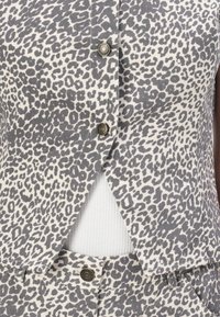 Blusa sem mangas em padrão de leopardo nas cores cinza e branco, com fecho de botão e tecido texturizado, apresentando um design em camadas sobre uma base branca com nervuras.