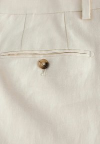 Baglommen på cremefarvede bukser med en enkelt brun knap og bæltestrop på struktureret stof.
