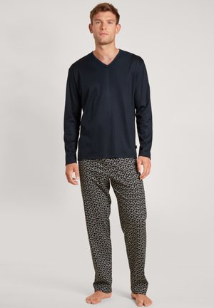 Man staat op blote voeten en draagt een donkerblauw V-hals shirt met lange mouwen en zwarte pyjamabroek met een kleine witte en rode bloemenprint.