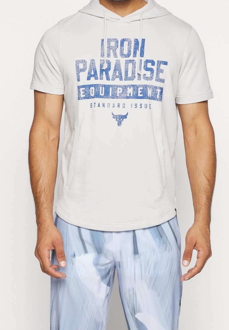 Homme portant un sweat à capuche blanc à manches courtes avec un texte bleu "IRON PARADISE EQUIPMENT" et un pantalon bleu clair à motifs, debout devant un fond uni.