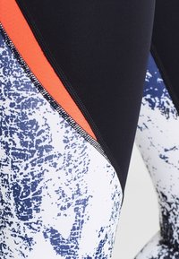 Närbild av sportleggings med svarta, orange och vita sektioner som har ett marinblått abstrakt mönster.