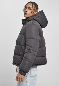 Dunkelgraue Pufferjacke mit Kapuze, gestepptem Design und elastischen Bündchen. Der Stoff hat eine glatte Textur ohne sichtbare Muster.